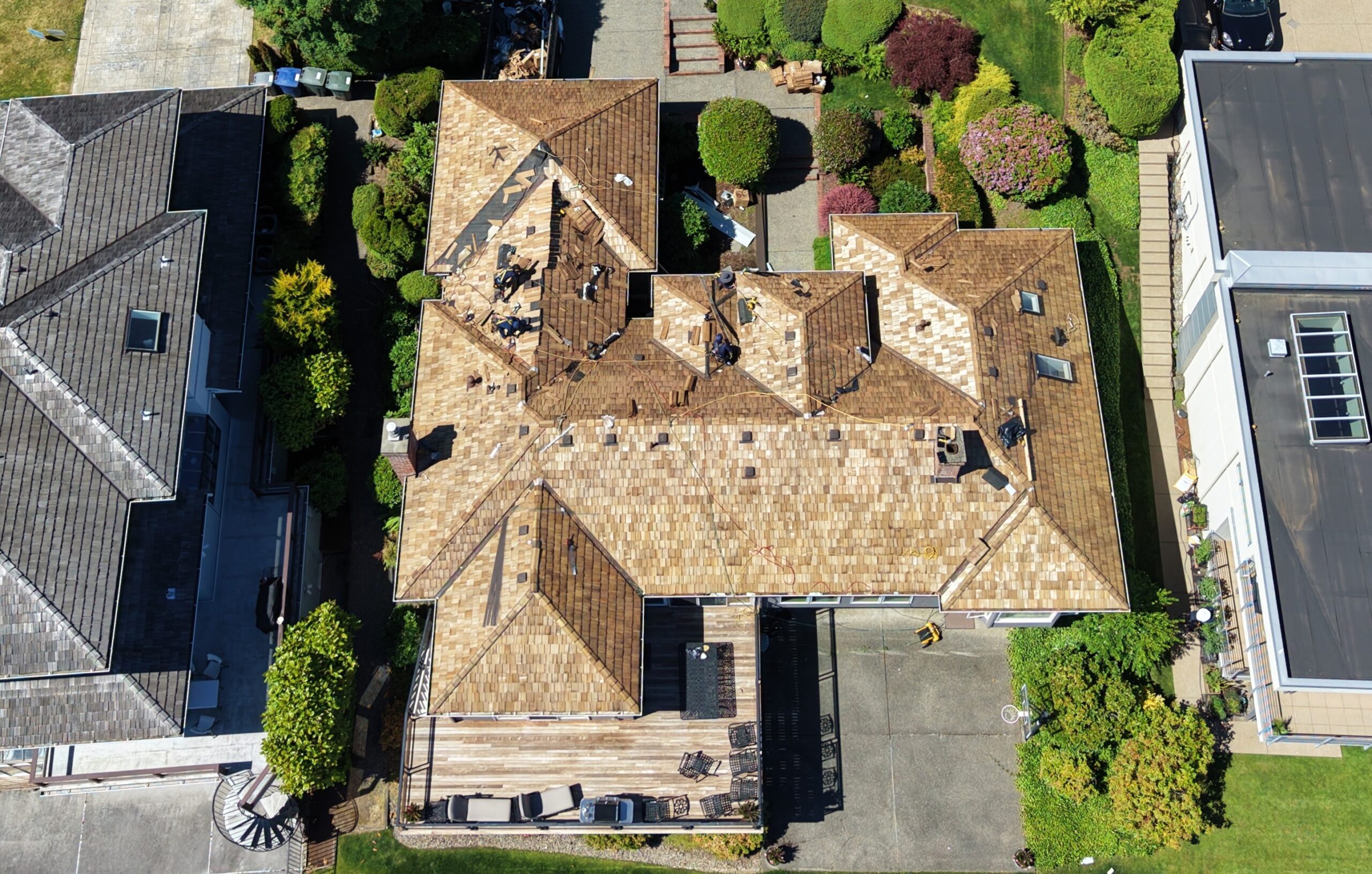 Aerial view of brown tiled roofs in a residential neighborhood; workers repair a central rooftop on a house nearby trees and driveways.