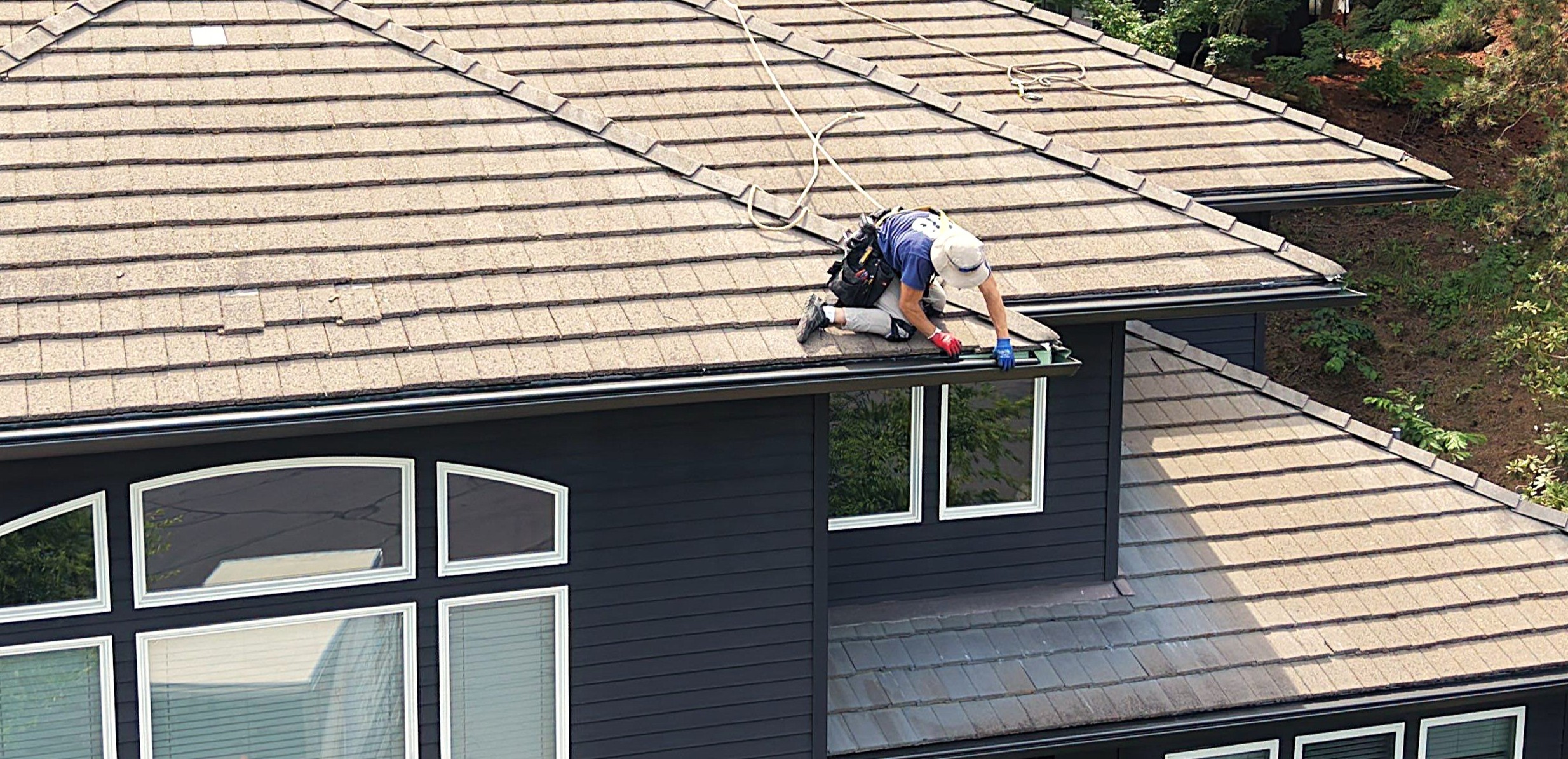 Worker on a sloped rooftop repairing shingles, wearing a helmet, gloves, and safety rope nearby.