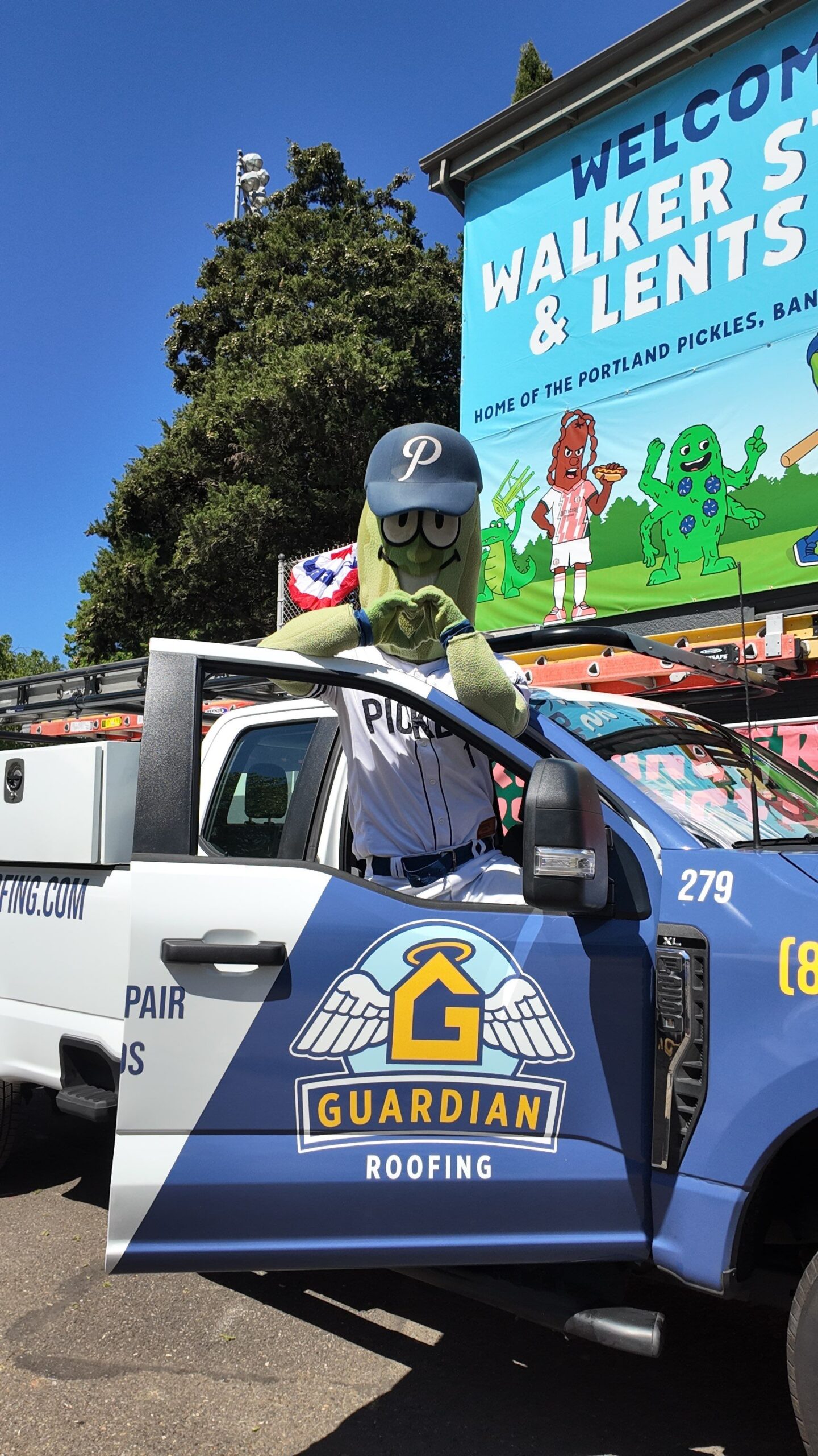 Mascot with glasses and a blue cap leaning on a Guardian Roofing truck at an outdoor event.