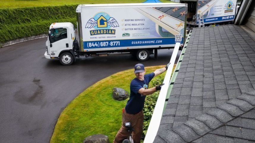 Worker on a ladder along a sloped roof edge near a Guardian Home service truck parked on the driveway, installing gutter/roof work in progress.