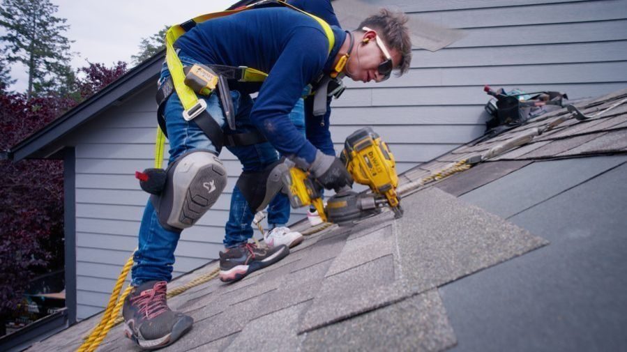 Worker on a sloped roof wearing safety gear, using a yellow cordless nail gun to install shingles.