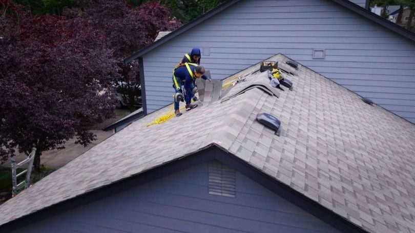 Two workers in safety gear patching gray roof shingles on a residential house from the ridge line