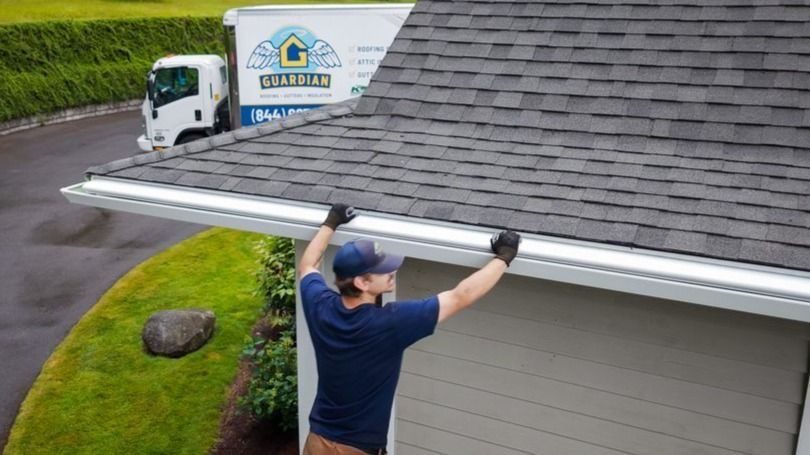 Man in a cap and gloves installs a white gutter along a gray shingled roof; a Guardian truck is in the driveway behind him.