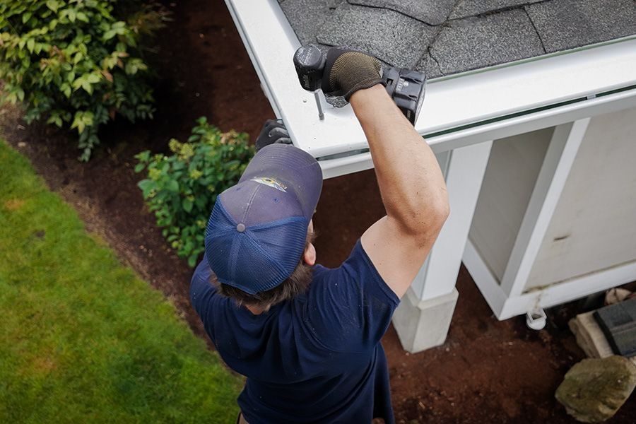 Construction worker on a ladder edges of a roof, fastening asphalt shingles with a power tool while wearing a blue cap and gloves.