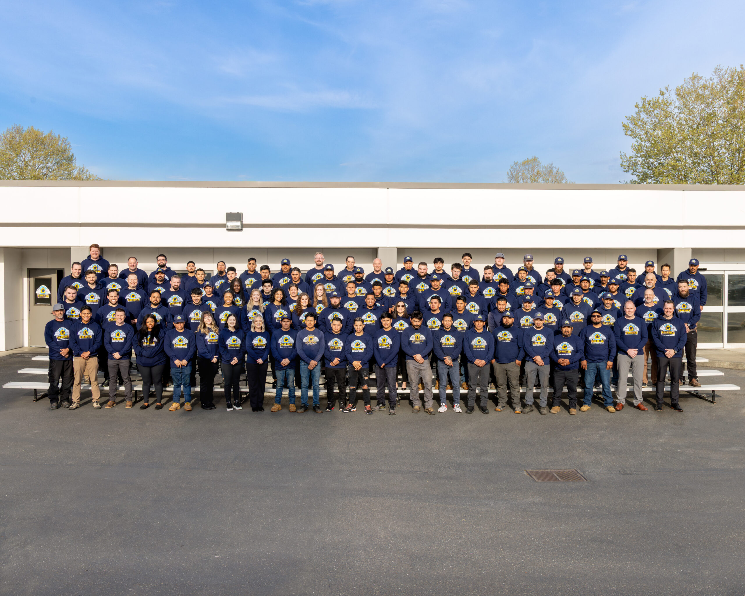 Large group of people in navy sweatshirts posing for a team photo outside a white, modern building against a blue sky.