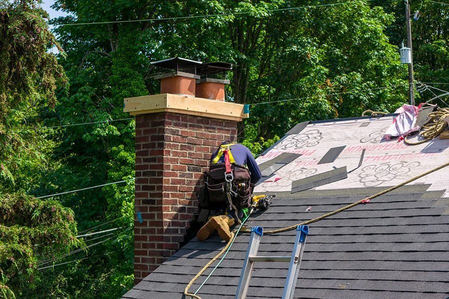 Roofer in safety gear on a sloped shingled roof, working near a brick chimney with a ladder nearby.